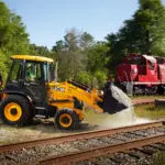 JCB 3CX Backhoe Loader carrying gravel and stones by railroad tracks.