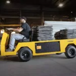 Worker driving Cushman Titan HD burden carrier through manufacturing facility with steel chained to the cargo deck.