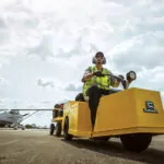Cushman Tug 8K tow tractor with attached trailer driving away from a grounded airplane.
