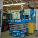 Genie GS-1930 slab scissor lift with workers in bucket repairing light.