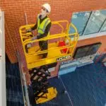 JCB 1930E scissor lift lifting an operator up next to a building.