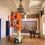 JLG 1230ES drivable vertical mast lift raising a worker to provide lighting maintenance in a large room.