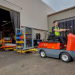 Taylor-Dunn C-426 AC heavy-duty sit-down tow tractor hauling trailer out of warehouse door.