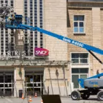 Workers repairing a clock on the side of a building in Genie S-60 DC Telescopic Boom Lift bucket.