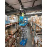 Genie GS-1932m aerial scissor lift in raised in warehouse aisle with workers in bucket.