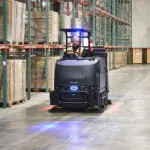 Nilfisk Advance CS7500 Sweeper Scrubber driving down warehouse aisle.